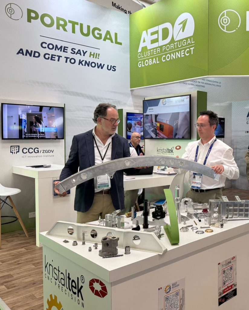 Stand de Kristaltek au Salon du Bourget 2025, intégré au pavillon du Portugal du cluster AED, présentant des composants CNC de haute précision pour l'industrie aérospatiale. Le propriétaire de l'entreprise, Joaquim Matos, et le CEO, Gonçalo Costa, présentent une pièce structurelle en aluminium aux visiteurs de l'événement.